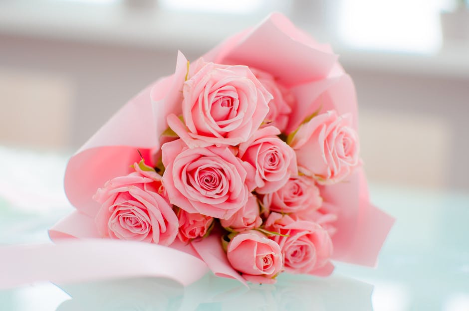 A bouquet of fresh pink roses arranged in a tight cluster, each with delicate, velvety petals in varying shades of light pink, wrapped in soft pink tissue paper with a hint of white. The bouquet is pl