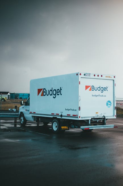 The image shows a white delivery truck with the 'Budget' logo and branding, parked outdoors on a wet surface under a cloudy sky. The truck is positioned in a slightly angled side view, revealing the c