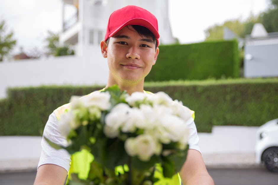 A young male flower delivery courier, wearing a red cap and a high-visibility vest, is smiling and holding out a bouquet of fresh white roses and green foliage towards the camera. The bouquet is arran