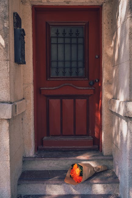 An exterior view of a red wooden front door with a black metal grate window and black door handle, set within a stone wall. At the base of the steps leading up to the door, there is a brown paper-wrap