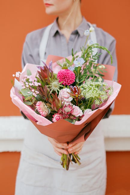 A hand holding a floral bouquet arranged with a variety of fresh flowers, including large white and purple chrysanthemums, a pink rose, and smaller blue and white blooms, all wrapped in elegant blue a