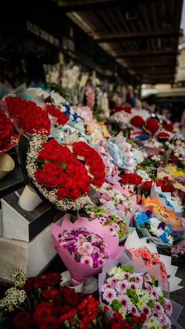 An outdoor flower stall displaying a vibrant array of floral arrangements and bouquets, primarily featuring red roses with lush green foliage, wrapped in white and pink paper, and accented with baby's