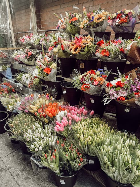 An indoor display of numerous flower bouquets arranged in black buckets and transparent wrapping, featuring a variety of fresh flowers including tulips in shades of pink, white, and red, alongside an 
