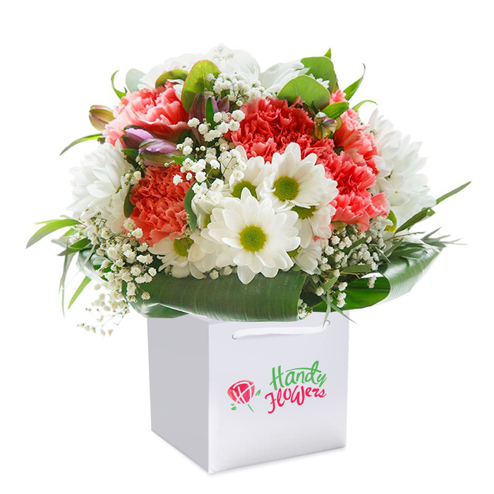 Set against a spotless white backdrop, this visually captivating floral arrangement embodies the artistry of Emerson Park's finest florists. Ruffled coral and soft peach carnations create a spirited foundation, their petals rich with texture and gentle energy. Clusters of crisp, white daisy-like chrysanthemums, each with inviting lime-green centers, evoke the cheerful ambience of Emerson Park's nearby gardens. Wisps of white baby's breath are carefully interwoven, imparting an ethereal quality that softens and balances the bold hues. Peek through the foliage to spot several unopened buds, tinged in purple and pink - their promise of bloom bringing a touch of intrigue, much like discovering a hidden path off Wych Elm Road. Deep green, glossy leaves envelop the base, framing the flowers in a polished collar that ties the design together. The bouquet sits upright within a smart, square white gift bag, topped with sturdy white ribbon handles for effortless transport. The composition speaks of freshness and quality, ideal for delivery to family, friends, or colleagues in Emerson Park - whether a birthday gesture, an anniversary celebration, or a simple moment of kindness. Each detail of this photographic presentation signals the florist's careful attention and deep appreciation for the local area's refined charm.