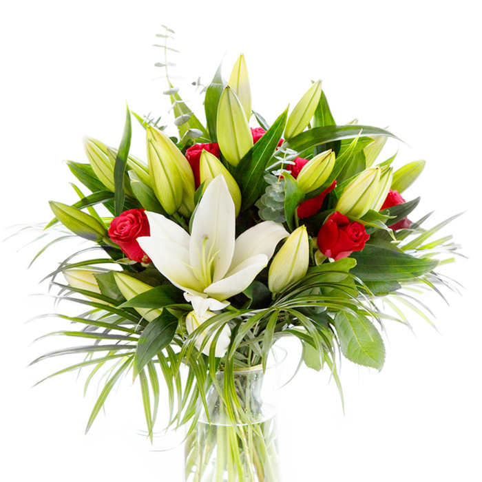 A classic, thoughtfully constructed bouquet photographed on a stark white background so each element reads with clarity: one prominent, fully open creamy-white Oriental lily takes center, its smooth petals slightly reflexed to display a pale yellow stamen and soft green pistil. Scattered throughout the arrangement are numerous lily buds in shades of lime green and pale yellow, hinting at successive blooms to come, while clusters of small, velvety red rosebuds add concentrated notes of passion and tradition. The greenery is abundant and layered for depth - glossy, structural dark green leaves frame the heart of the bunch, elongated palm-like fronds cascade outward providing movement and silhouette, and silvery-green eucalyptus stems with their round leaves introduce a subtle, cool texture. The flowers are gathered in a clear cylindrical glass vase so the fresh green stems and clear water are visible, reinforcing a sense of freshness and straightforward elegance. The bright studio lighting highlights textures from the lily's waxy surface to the rose's velvet and the eucalyptus's matte silver, creating a tactile impression even without scent. This arrangement is suitable for many occasions: romantic gestures, birthdays, or a tasteful sympathy tribute, and it carries the quiet professionalism of a local Emerson Park florist who knows how to balance purity and passion for customers across Hornchurch, Harold Wood and nearby neighbourhoods.