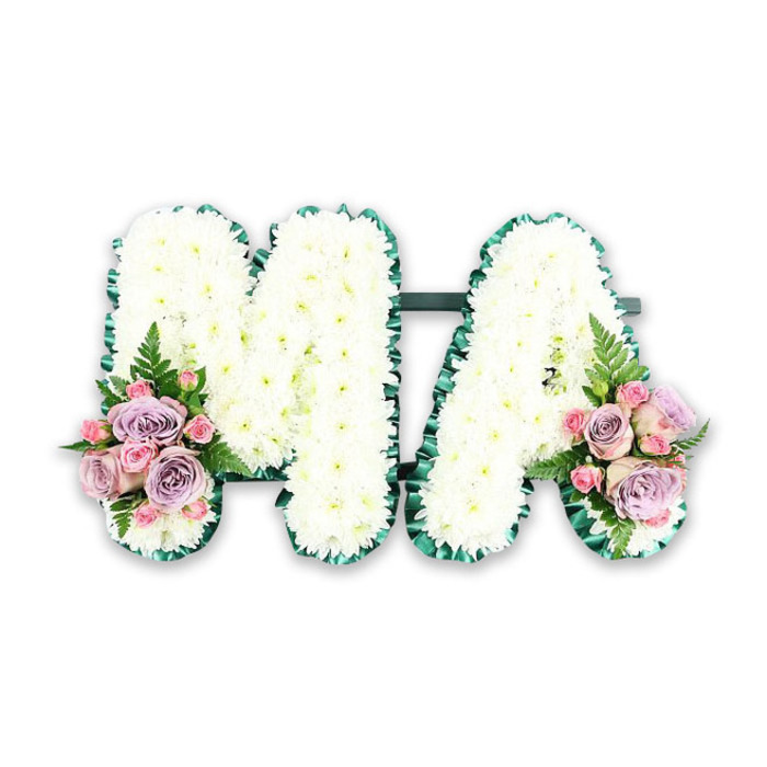 This image shows an elegant funeral letter tribute spelling out MA, presented against a bright white backdrop so that every flower and leaf can be fully appreciated. The design is meticulously structured: the letters stand side by side, joined at the base by a subtle horizontal bar that keeps the tribute secure for display on an easel or coffin during a service in or around Emerson Park. Each stroke of the M and the A is densely packed with crisp white chrysanthemums, the small, rounded blooms pressed so closely together that they create a continuous, cushion-like surface. The flowers appear freshly cut, their petals slightly ruffled yet tidy, giving the letters a soft but well-defined appearance. Along the outer edges, a deep emerald green border-likely satin ribbon or compact greenery-gently ripples in a pleated pattern, tracing the contours of each letter. This dark green outline provides a striking contrast to the whiteness of the blooms and ensures the word MA remains legible from a distance, whether placed at the front of a church in Hornchurch or at a graveside near Emerson Park. At the lower left of the M and the lower right of the A, two matching accent arrangements bring a touch of colour. Each cluster contains lilac or dusty purple roses, pale blush-pink roses and tight pink spray rose buds, all tucked closely together to form rounded posies. Soft fern fronds and a few sprigs of fresh foliage extend beyond the blooms, adding shape, texture and a natural, garden-inspired feel. This carefully balanced composition makes the tribute especially fitting for remembering a beloved mother or maternal figure, expressing tenderness, gratitude and enduring love through its thoughtful use of colour and texture.