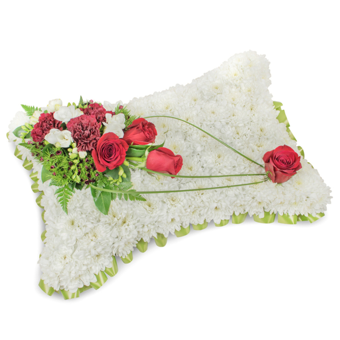 Arranged as a traditional memorial pillow, this rectangular floral tribute is a study in contrast and calm: the entire surface is covered with a dense layer of pristine white chrysanthemum blooms, each tiny head contributing to a plush, fleece-like texture that communicates rest and purity. Around the slightly rounded edges, narrow glimpses of a pale green ribbon or delicate foliage edge the form, giving a neat, respectful border. At the upper left a composed spray of deep red flowers-several roses from tight bud to full bloom alongside frilly, dark burgundy carnations-creates a bold focal cluster. Intermingled are small white accent blooms, similar to freesias or miniature chrysanthemums, which add lightness and a faint floral sweetness. Layers of lush greenery, including fine fern fronds and broader leaves, tuck under and behind the red cluster, providing depth and a living green framework. From this main cluster, three elegant strands of bright green bear-grass or slender decorative foliage arc diagonally across the pillow toward the bottom right, producing a graceful line that connects the corners. The terminal point of that sweep is a single, immaculate red rose positioned on the lower right - a solitary emblem of love and remembrance, delicately held by the curving stems. The overall composition is understated and carefully balanced, with soft, diffused lighting that highlights petal texture and the quietly rich reds against the white field. This type of cushion is often chosen for funeral services and memorial displays in local churches and chapels near Emerson Park and the surrounding Hornchurch community as a timeless, comforting expression of sympathy.