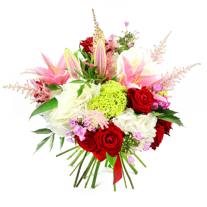 This hand-tied bouquet, photographed against a bright white background, showcases a harmonious blend of romantic hues and luxurious textures, reminiscent of a carefully considered floral display in a stylish Emerson Park lounge. Central to the arrangement are classic deep red roses, their velvety, tightly layered petals forming perfect rosettes that exude passion and sophistication. Interspersed among these are tall pink lilies, some fully open with broad, curved petals that reveal pale, delicately marked throats and softly dusted stamens, while others remain as smooth buds, pale green at the tips, promising future blossoms and fragrance. The roses and lilies are cradled by generous clusters of ruffled white flowers-similar to hydrangeas or ornamental carnations-that create a plush, airy base, giving the bouquet a sense of abundant volume and serene lightness. Near the centre, a striking, rounded burst of lime green dianthus or Green Trick carnation adds a vivid, mossy texture, providing a contemporary focal point that contrasts beautifully with the smoother petals around it. Feathery sprays of light pink astilbe rise and arch gracefully from various points, lending a delicate, almost ethereal quality that softens the outer edges of the design. Tucked discretely among the more prominent blooms, small sprigs of fuchsia and lavender filler flowers add nuanced touches of richer colour, encouraging closer visual exploration. Dark, glossy foliage is woven throughout, framing the brighter hues and grounding the composition in lush greenery. At the bottom, the stems are neatly trimmed and held tightly together with a slim red satin ribbon, clearly identifying the bouquet as expertly hand-tied and ready to present. The overall impression is of joyful elegance and considered luxury, perfect for celebrating a special anniversary, marking a birthday, or sending a warm, thoughtful message to friends and family across Emerson Park, from the quiet roads off Nelmes Way to the homes near Hornchurch Country Park.