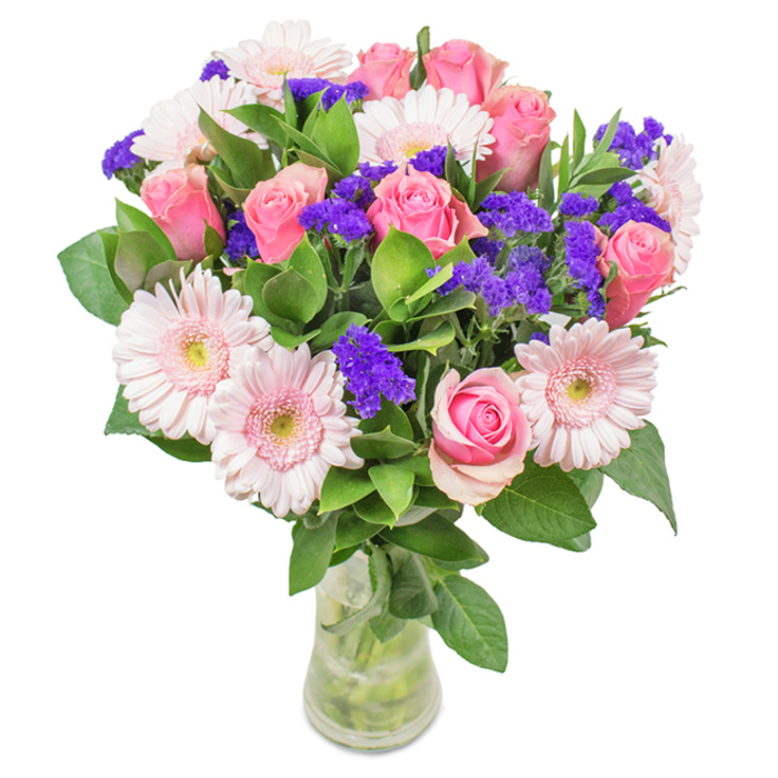 An artfully arranged floral bouquet takes pride of place in a clear glass vase against a bright white background, echoing the classic charm of Emerson Park interiors. A focal point of seven to nine pink roses, some tightly closed, others gently unfurling, showcases traditional elegance and subtle romance. They are delightfully complemented by five to seven pale pink gerbera daisies, their round, ray-like petals and soft white cores providing an inviting sense of innocence and cheer. Threaded through the arrangement are dynamic purple statice, their vivid hue adding texture and depth, much like the colourful gardens hidden amongst Emerson Park lanes. A base of lush, glossy green leaves provides eye-catching contrast and a fresh, abundant feel reminiscent of spring in local parks. Crystal-clear water and stems visible through the vase reinforce the bouquet's freshness. Perfect for occasions such as birthdays, anniversaries, Mother's Day, or for decorating a bright Emerson Park kitchen, this bouquet projects feelings of joy, celebration, and sincere affection-an inviting gesture from the heart of the community.